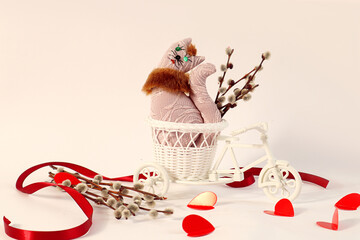  Holiday card: a soft toy - a pastel cat on a decorative white bicycle, willow branches, red hearts, a scarlet ribbon on a light background, space for text