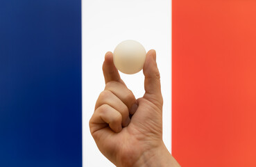 The hand holds a table tennis white ball in front of France flag.