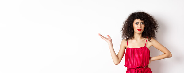 Upset pretty woman with curly hair and red dress, raising hand and look confused, frowning...
