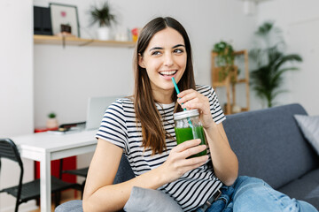 Happy young woman drinking healthy green juice sitting on sofa at home. Pretty millennial girl...
