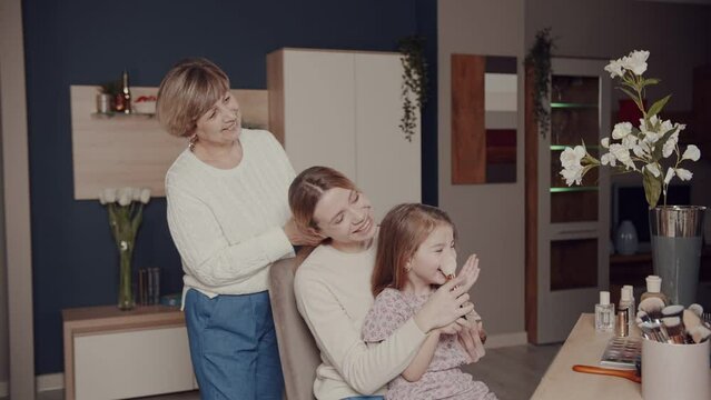 Three beautiful women from different generations smile and laugh while looking in the mirror while applying blush on their daughter's cheeks. Grandma combs Mom's hair. Parental care. Happy family.