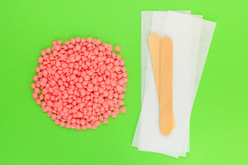 Beautiful pink depilatory wax granules, strips for depilation and wooden spatulas on green background. Epilation, depilation, unwanted hair removal. Top view.