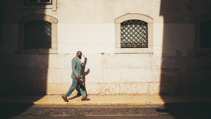 A side view of an dapper bald bearded black man entrepreneur in a green tailored summer suit passing by an illuminated part of the street next to the wall in between two shadows, putting on sunglasses