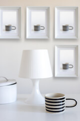 A black and white striped mug, a lamp with a shade and a cookie box on a white table. On the wall are photographs of this mug in three frames. Vertical frame. Space for text.