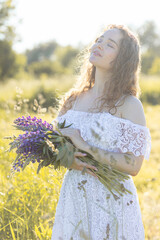 Fototapeta na wymiar Close up portrait of happy smiling beautiful young girl in a white dress with curly hair, straw hat, with picnic and bouquet of purple wild flowers on a meadow. Summertime, golden hour, sunset