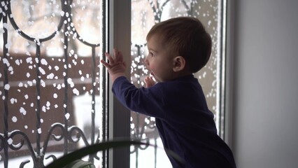 A small child stands on the windowsill and looks out the window. A child knocks on glass in winter.