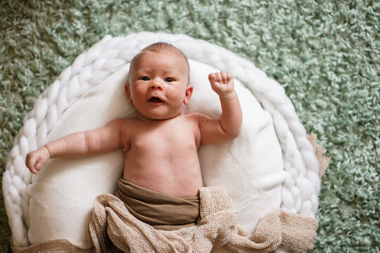 happy cheering naked newborn baby lying on a white round bed with arms up and fists looking into the camera as a winner shouting yay hooray