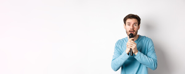 Confused young man looking nervously at camera while singing karaoke, holding microphone, standing...