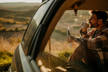 Fototapeta na wymiar A man sitting in the open car with a coffee thermos flask and smiling. A young man having a coffee break during his road trip.
