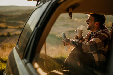 A man sitting in the open car with a coffee thermos flask and smiling. A young man having a coffee...