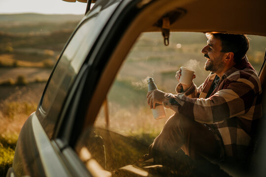A man sitting in the open car with a coffee thermos flask and smiling. A young man having a coffee break during his road trip.