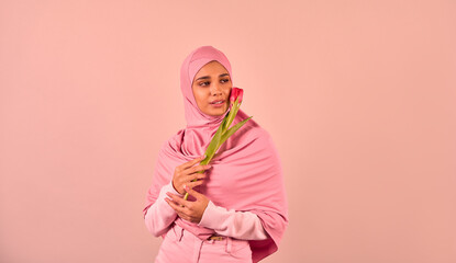  Young beautiful Muslim woman wearing soft pink hijab and clothes holding tulip and looking away while standing near beige background.