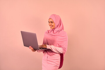   Beautiful modern young Muslim woman wearing pink hijab and stylish clothes holding laptop while...