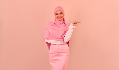   Stylishly dressed modern Muslim woman standing near a beige background and pointing with her...