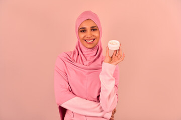  Beautiful young Muslim woman standing on beige background, showing face cream and smiling. Skin...