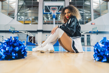 Curly-haired cheerleader in black and white uniform sitting on basketball court. Blue shiny...