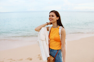 Woman smile with teeth after swimming in the ocean with a backpack in a wet yellow tank top and denim shorts walks along the beach, summer vacation on an island by the ocean in Bali sunset