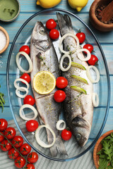 Fototapeta na wymiar Glass baking tray with sea bass fish and ingredients on light blue wooden table, flat lay