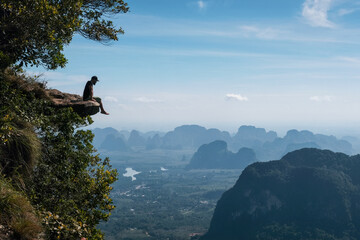 A tourist sits on the edge of a cliff overhanging a abyss on sunny day. Dragon's Crest (Khao Ngon Nak) Viewpoint, Krabi Province, Thailand.