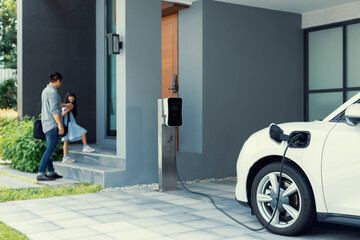 Fototapeta Focus progressive electric vehicle recharging at home charging station using clean and renewable energy with blurred father and daughter walking in background for future renewable energy concept. obraz