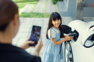 Progressive mother using smartphone take focus photo of daughter recharge EV car. Image of asian...