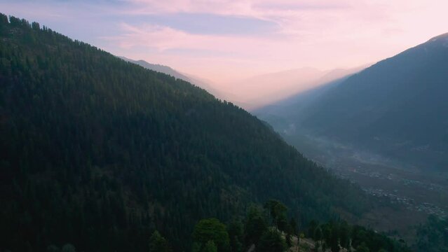 aerial drone shot sideways towards layers of mountains of himalaya range with tree covered hills and pink sky with rays showing manali town from hamta village a tourist spot
