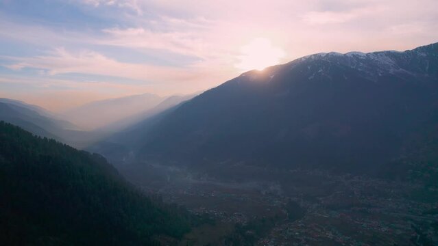 aerial drone shot flying towards layers of mountains of himalaya range with tree covered hills and pink sky with sun just behind mountain and rays showing manali town from hamta village a tourist spot