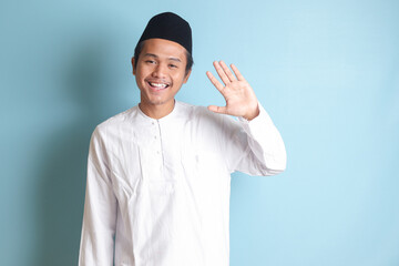Portrait of Asian muslim man in white koko shirt with skullcap shirt saying hi, waving hand at camera, greeting person with happy smile. Isolated image on blue background - obrazy, fototapety, plakaty