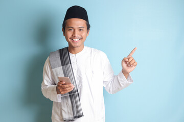 Portrait of young Asian muslim man holding mobile phone with smiling expression on face while pointing finger to the side. Isolated image on blue background