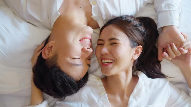 Top view of true love young asia couple lying down kiss wife face cheek on cozy bed. Relax awake morning fall in love of just married sweet lover asian people joy laugh toothy smile at home bedroom.