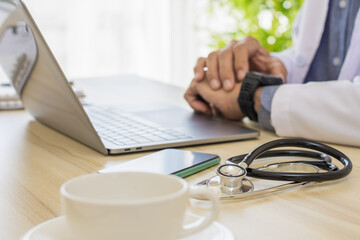 Male doctor looking at smart watch while work on laptop computer with medical stethoscope and mobile smartphone on the desk at clinic or hospital.