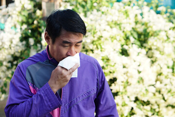 Asian man catch a cold, holding white tissue paper to wipe his nose, standing beside blooming flowers at the park. Concept : Pollen allergy symptoms, sick, runny nose, sneeze. Health problems.  - obrazy, fototapety, plakaty