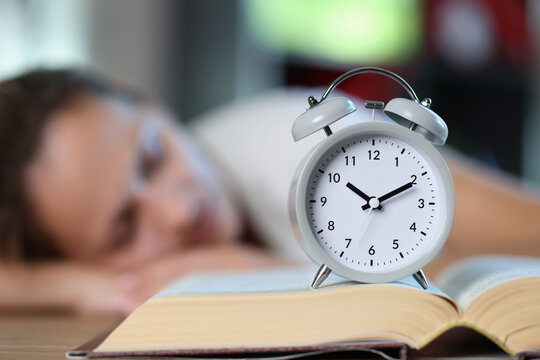 Open Book And Alarm Clock Close Up, Sleeping Woman In Background.