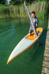 Fototapeta na wymiar Junger Mann beim Trendsport Stehpaddeln, auf dem Board kniend beim Ablegen vom Steg am See