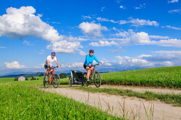 Eltern mit Nachwuchs im Buggy unterwegs mit dem Mountainbike, die Voralpen-Landschaft im Allgäu...