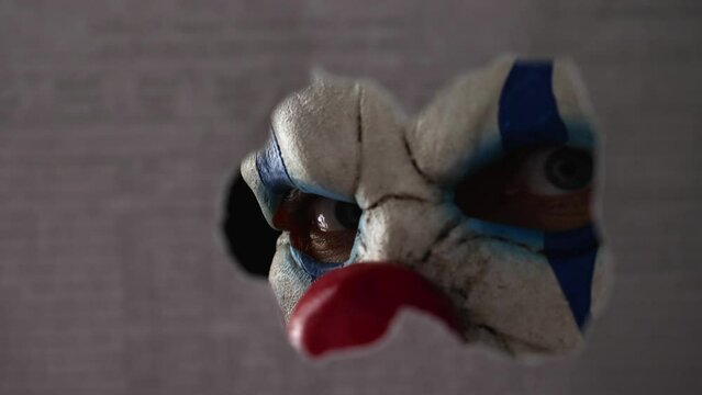 A scary clown face peeking through a hole in the partition. Close-up. The face of a man wearing a creepy clown mask peering at his victim through the hole. The creepy clown maniac