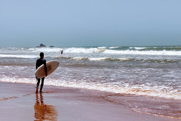 Unknown man in a wetsuit standing with the surfboard in his hands along the Atlantic coast. Essaouira, Morocco.