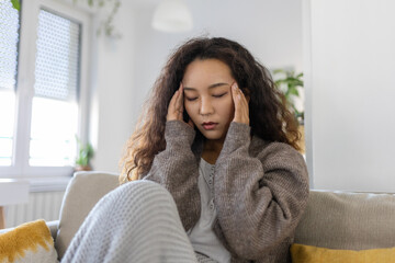 Portrait of a young Asian woman sitting on the couch at home with a headache and pain. Beautiful woman suffering from chronic daily headaches. Sad woman holding her head because sinus pain