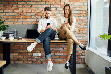 Two businesspeople sitting on desk at modern startup office using smartphones