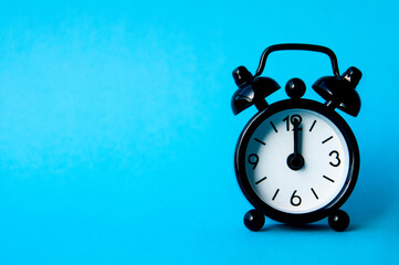 Black alarm clock isolated on blue background. The clock set at 12pm. Copy space and time concept