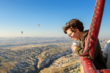 Young woman in hot air balloon basket, looking at balloons and beautiful landscape of Goreme,...