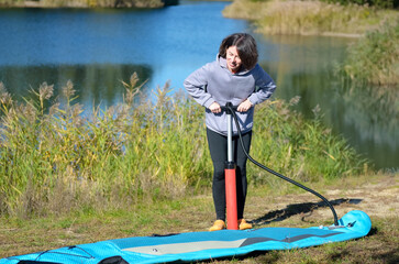Active woman inflates SUP board with manual pump, beautiful lake and nature on background, stand up...