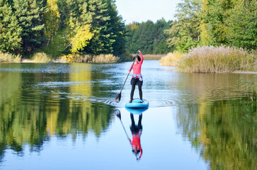 Fototapeta na wymiar Active woman paddling SUP board on beautiful lake, autumn forest landscape and nature on background, stand up paddling water adventure outdoors, sport and healthy lifestyle concept 