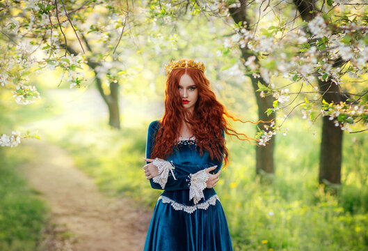 Redhead fantasy woman queen. Blue long velvet dress vintage old style clothing. Red curly hair flying waving in wind. Summer nature green flowering trees garden. Girl Princess beauty face. art photo
