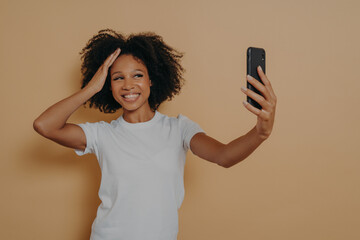 Happy charming dark skinned female making photo selfie on modern smartphone against beige wall