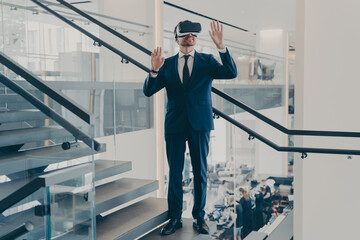 Businessman in VR headset stands on staircase in office lobby with hands up in air
