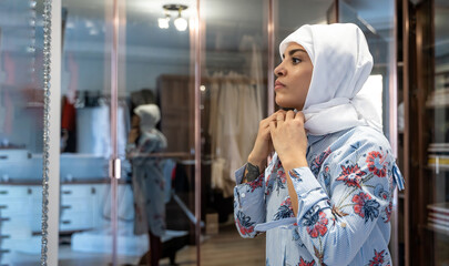 Portrait of middle-aged muslim woman wearing put on hijab headscarf in dressing room at home looking at mirror.Side view face of cheerful woman covered with headscarf. Casual islamic girl. Copy space