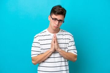 Young handsome Brazilian man isolated on blue background keeps palm together. Person asks for...