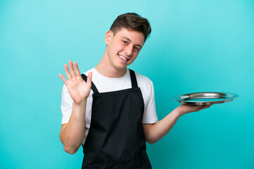 Young waitress with tray isolated on blue background saluting with hand with happy expression © luismolinero