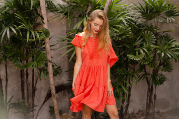 Happy smiling blond woman in elegant red dress posing   in stylish bali style resort  during tropical vacation.